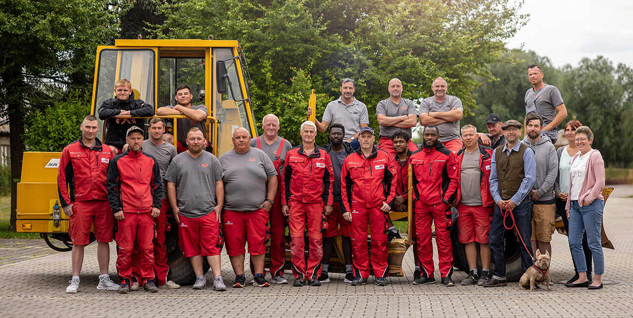 Teamfoto: 23 Mitarbeiter von SK Sportbau in roter Arbeitskleidung vor gelbem Bagger